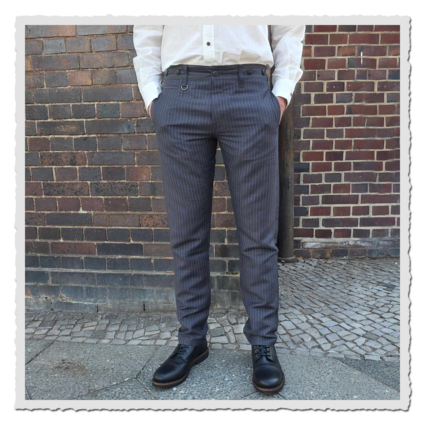 Vorderansicht einer grauen Nadelstreifenhose am Model vor gemauerter Backsteinwand im Außenbereich.